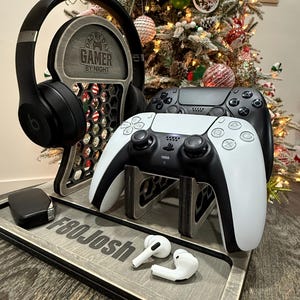 May include: A wooden gaming console stand with a gray finish. It features a headphone holder with the text "Dad by Day Gamer by Night" and slots for two controllers and a charging pad. The stand also has a personalized inscription "F80Josh".