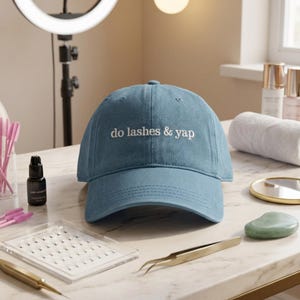 May include: A blue denim baseball cap featuring the white embroidered text "do lashes & yap" centred on the front panel. The cap has a structured crown, a curved brim, and stitched eyelets, presented as a standalone accessory item.