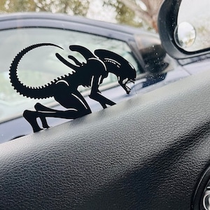 Peut inclure: Silhouette noire d'une créature extraterrestre rampant sur le tableau de bord d'une voiture noire.