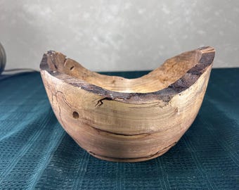 Könnte beinhalten: Eine handgefertigte Holzschale mit natürlicher Waldkante und dunklen, unregelmäßigen Rindenstrukturen. Die Schale zeigt eine hellbraune Holzmaserung mit markanten dunkelbraunen Streifen, natürlichen Astlöchern und einer glatten, geschwungenen Innenfläche.