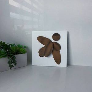 Puede incluir: Una escultura de madera marrón de un ángel estilizado con alas, montada sobre un fondo cuadrado blanco.