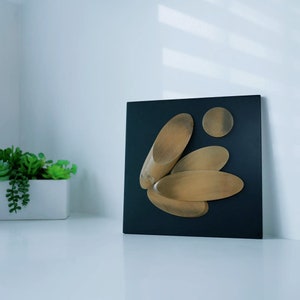 Puede incluir: Un lienzo cuadrado negro con cinco formas de madera en tonos marrones, dispuestas en un racimo. Las formas son orgánicas y se asemejan a hojas o pétalos.