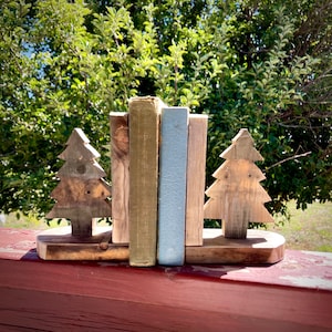 May include: Wooden bookends in the shape of Christmas trees, holding vintage books. The bookends are made of light-colored wood and sit on a wooden base. The books have various colors and textures, creating a rustic, natural aesthetic.