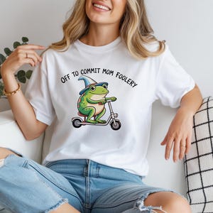 May include: White t-shirt with the text "Off to Commit Mom Foolery." The shirt features a cartoon frog wearing a wizard hat and riding a scooter. The frog is green with red spots.
