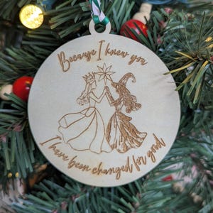 May include: A round wooden Christmas ornament with the engraved text "Because I knew you, I have been changed for good." The ornament features illustrations of two figures. It hangs from a green and white ribbon, set against a Christmas tree.