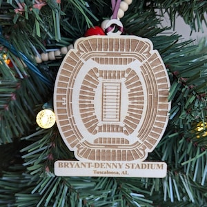 May include: A wooden ornament shaped like Bryant-Denny Stadium in Tuscaloosa, Alabama. The ornament is brown and features a detailed outline of the stadium.