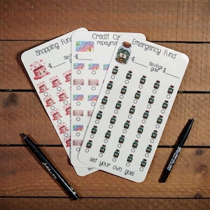 May include: Three white budget tracker cards with different designs. One card has shopping bags, another has a credit card graphic, and the third has a jar of money. Two black pens are also in the image. The cards have the text "Shopping Fund", "Credit Card repayment", and "Emergency Fund".