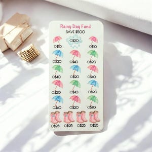 Peut inclure: Une carte blanche avec le texte "Rainy Day Fund SAVE 500$" présente des illustrations à l'aquarelle de parapluies, de nuages de pluie et de bottes de pluie. La carte est conçue pour suivre l'épargne, avec des montants allant de 10 $ à 40 $.