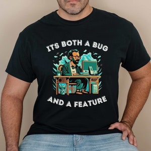 May include: A black t-shirt with a white graphic of a person sitting at a desk with a computer. The text on the shirt reads "ITS BOTH A BUG AND A FEATURE".