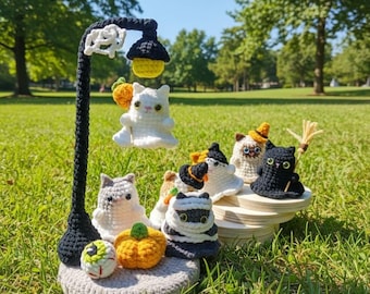 May include: A whimsical crochet Halloween display featuring a black lamp post with a yellow light, a white cat ghost, and various crochet cats, pumpkins, and an eyeball. The display is set on a gray crocheted base and a wooden platform.