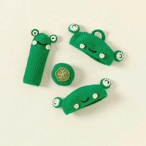 Può includere: Quattro giocattoli di rana in crochet verde. Due sono fasce per capelli, uno è un piccolo giocattolo rotondo e uno è un giocattolo lungo e sottile. Tutti hanno occhi bianchi e una bocca nera.