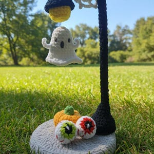 May include: A crochet Halloween decoration. A black lamp post with a yellow lamp and a white ghost. The base holds a pumpkin and two eyeball ornaments, all on a grey crocheted base. The scene is set outdoors on a grassy lawn.
