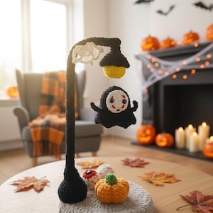 Puede incluir: Un farol de ganchillo con temática de Halloween hecho a mano, con una base negra, una luz amarilla y una figura de fantasma negro flotante. La base presenta una pequeña calabaza naranja y otros artículos de ganchillo con temática de Halloween.