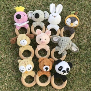Op de afbeelding: Een verzameling gehaakte babyrammelaars in verschillende dierenvormen, waaronder een koala, konijn, varken, olifant, panda en beer. Elke rammelaar heeft een houten ring en is gemaakt in zachte pastelkleuren.