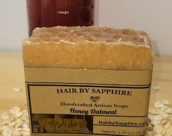 Puede incluir: Una barra de jabón artesanal con una etiqueta marrón que dice "Hair By Sapphire Handcrafted Artisan Soaps Honey Oatmeal HairbySapphire.com Approx. 4.18-5.12 Oz". El jabón está sobre una superficie con avena esparcida alrededor. Una botella de miel está en el fondo.