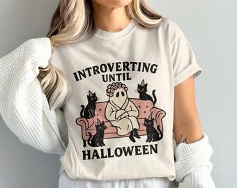 Puede incluir: Camiseta color crema con la frase "INTROVERTING UNTIL HALLOWEEN". El diseño presenta un personaje fantasma en un sofá rosa rodeado de gatos negros con sombreros de fiesta. Un diseño divertido y espeluznante para Halloween.