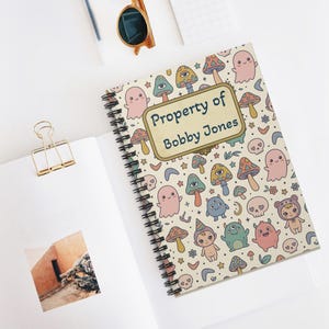 May include: Spiral-bound notebook with a whimsical pattern of mushrooms, ghosts, and other cute characters. The cover features the text "Property of Bobby Jones" in a decorative frame. The notebook is open to a blank page.