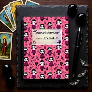 Puede incluir: Un cuaderno con espiral de color rosa con una cubierta que presenta un patrón repetido de figuras de dibujos animados. El cuaderno tiene el texto "UNSUPERVISED THOUGHTS" y "Dr. Beatles" en la portada. También hay cartas del tarot, esferas negras, un bolígrafo y amuletos con forma de murciélago.
