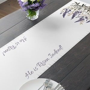 May include: A white table runner featuring a botanical illustration of purple lavender sprigs at both ends. The text "He is Risen!" and "He is Risen Indeed!" is printed in a dark script font along the center of the fabric runner.