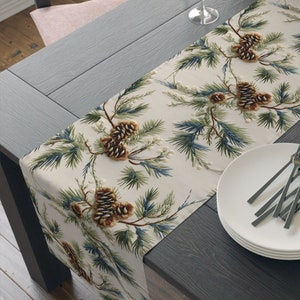 May include: A white table runner with a pine cone and evergreen branch design. The runner is on a dark wood table with a stack of white plates and silverware.