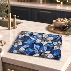 May include: A rectangular glass cutting board featuring a vibrant stained glass pattern of blue and white poinsettia flowers with gold-toned outlines. The design covers the entire surface of the board, showcasing intricate floral petals and leaves.