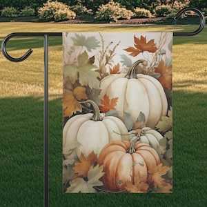 May include: A garden flag featuring a watercolor illustration of pumpkins and fall foliage. The pumpkins are white and orange, surrounded by green, orange, and brown leaves. The flag is displayed on a black metal stand.