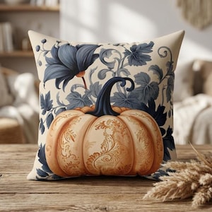 May include: A square throw pillow featuring a cream-colored background with a central orange pumpkin illustration. The pumpkin has intricate gold-toned filigree patterns and a dark blue stem. Dark blue floral and leafy vines surround the pumpkin.