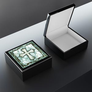 K&ouml;nnte beinhalten: Eine schwarze Holzkiste mit einem Scharnierdeckel. Der Deckel ist ge&ouml;ffnet und das Innere ist mit wei&szlig;em Filz ausgekleidet. Die Kiste hat eine dekorative Oberseite mit einem Buntglasdesign eines wei&szlig;en Kreuzes auf blauem und gr&uuml;nem Hintergrund.