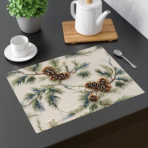 May include: A white placemat with a pine cone and evergreen branch design. The placemat has a cream background with brown pine cones and green and blue evergreen branches.