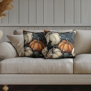May include: Two square throw pillows featuring a repeating pattern of orange and cream-colored pumpkins set against a dark background with dark blue and green foliage. The pillows have a smooth texture and are displayed on a light-colored sofa.