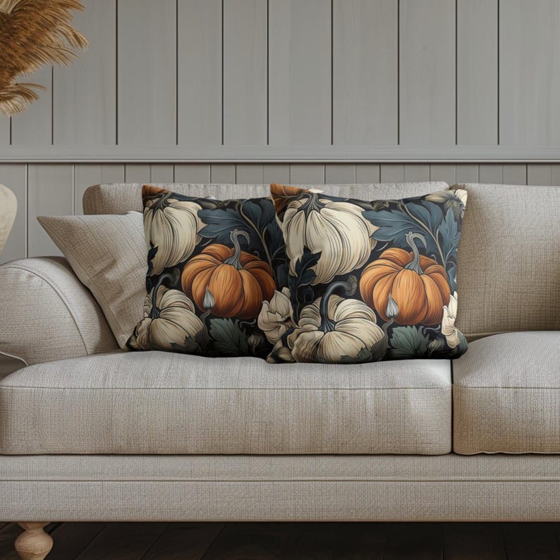 May include: Two square throw pillows featuring a repeating pattern of orange and cream-colored pumpkins set against a dark background with dark blue and green foliage. The pillows have a smooth texture and are displayed on a light-colored sofa.