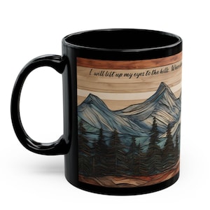 May include: A black ceramic mug featuring a wrap-around graphic of a mountain range and pine forest with a wood-grain texture background. The text "I will lift up my eyes to the hills" is printed in black script near the top of the mountain illustration.