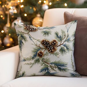 May include: A square throw pillow featuring an off-white fabric base with an embroidered pattern of green and blue pine needles, brown pinecones, and small white berries. The embroidery has a raised, textured appearance across the surface.