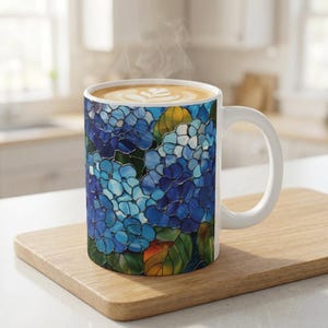 以下が含まれることがあります： 青いアジサイの花と緑や黄色の葉が描かれたステンドグラス風の模様が特徴の白いセラミック製マグカップ。マグカップにはラテアートが施された温かい飲み物が入っており、カップの上部からは湯気が立ち上っています。