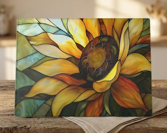 May include: A rectangular glass cutting board featuring a vibrant stained glass style sunflower design. The artwork displays overlapping petals in shades of yellow, orange, and deep brown, with accents of teal and green throughout the composition.