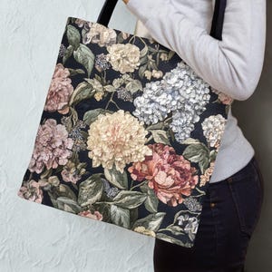 May include: A tote bag with a dark background and a floral pattern featuring large, colorful flowers in shades of pink, cream, and white. The bag has black handles and a rectangular shape, suitable for carrying items.