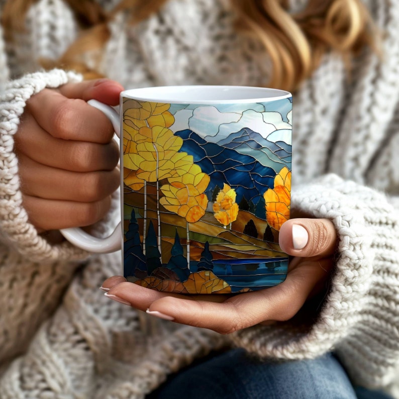 May include: White ceramic mug with a stained glass design of a mountain landscape with yellow trees and a blue lake.