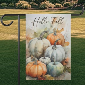 May include: A rectangular garden flag featuring a watercolor illustration of various pumpkins in shades of orange, white, and blue-grey. The text Hello Fall is printed in a black cursive script at the top. The flag hangs from a black metal stand.