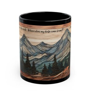 May include: A black ceramic mug featuring a wrap-around graphic of layered mountain peaks and pine trees over a wood-grain background. The text "I lift up my eyes to the hills. Where does my help come from?" is printed in black script near the top rim.