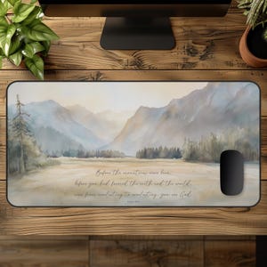 May include: A rectangular mousepad featuring a watercolor landscape of mountains and a valley in soft blues and greens. The text "Before the mountains were born..." is printed in a script font. A black computer mouse is on the right.