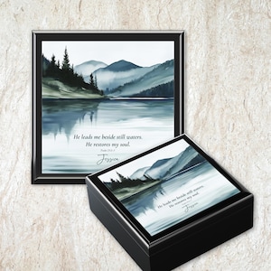 May include: A framed watercolor print and a matching black box featuring a serene landscape of mountains and water in shades of blue and green. The text "He leads me beside still waters. He restores my soul. Jessica" is displayed on both items.