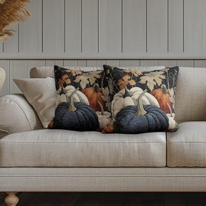 May include: Two square throw pillows featuring a dark background with a repeating pattern of orange, white, and dark blue pumpkins, along with light-colored autumn leaves. The pillows have a quilted texture and are placed side by side on a sofa.
