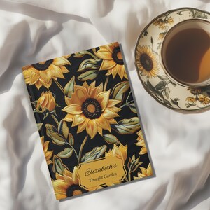 May include: A hardcover journal with a sunflower design and the text "Elizabeth's Thought Garden" on a gold label. The journal is next to a teacup and saucer with a sunflower motif. The background is a white fabric.