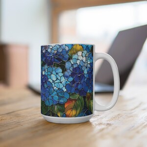 以下が含まれることがあります： 青いアジサイの花と緑の葉の鮮やかなステンドグラス模様が施された白いセラミック製のマグカップ。デザインはマグカップの外側全体を覆っており、右側に湾曲したハンドルと滑らかな円筒形の本体を備えています。