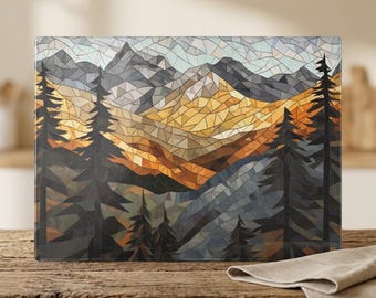 May include: A rectangular glass cutting board featuring a mosaic-style graphic of a mountain landscape. The design displays dark grey pine trees in the foreground against a backdrop of golden-yellow and grey mountain peaks under a pale sky.