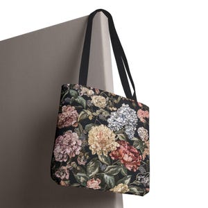 May include: A tote bag with a floral design. The bag has a dark background with flowers in pink, yellow, and white. It has black handles and is hanging against a neutral wall. The bag's design is reminiscent of a tapestry.