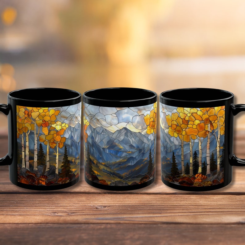 May include: Three black ceramic mugs with a stained glass design of a mountain landscape with trees. The design features a blue mountain range with a yellow and orange sunset sky.