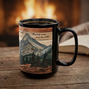May include: A black ceramic mug featuring a graphic illustration of a mountain range and pine forest. The text reads, My help comes from the LORD. Psalm 121:1-2. The design is set against a wood-grain background with a dark handle.