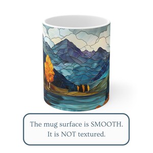 May include: A white ceramic mug with a stained glass design of a mountain range, a lake, and a tree with orange leaves. The mug surface is smooth.