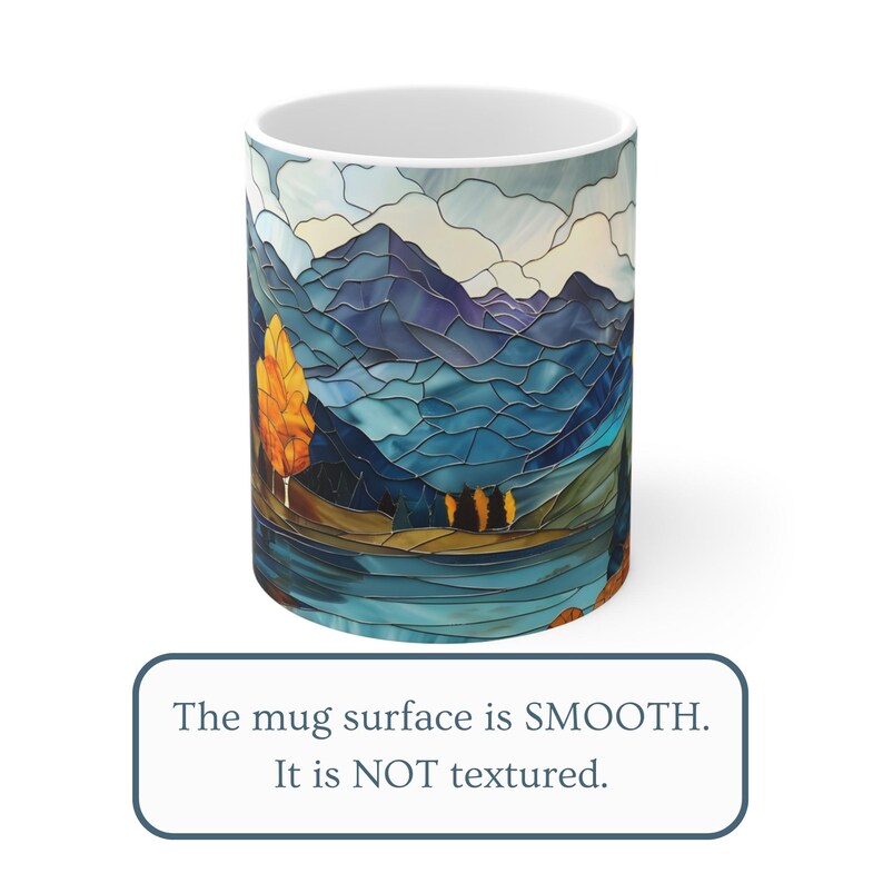 May include: A white ceramic mug with a stained glass design of a mountain range, a lake, and a tree with orange leaves. The mug surface is smooth.